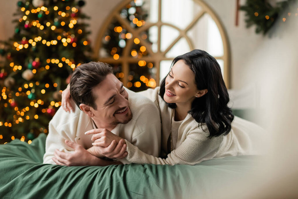 portrét veselého páru ležícího spolu na posteli v blízkosti zdobené jasný vánoční stromeček se světly - Fotografie, Obrázek