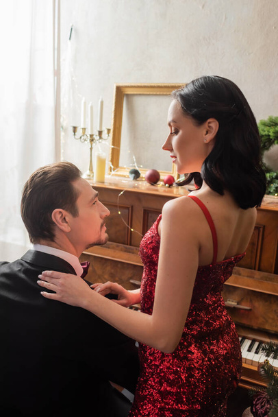rijk paar, aantrekkelijke vrouw in rode jurk staan naast echtgenoot in smoking en piano - Foto, afbeelding