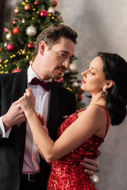 schöner Mann im Anzug hält Hand einer hübschen Frau im roten eleganten Kleid in der Nähe des Weihnachtsbaums - Foto, Bild
