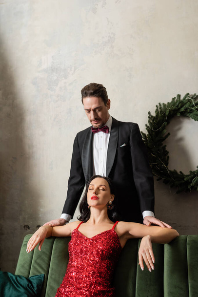 Красивый джентльмен в смокинге смотрит на красивую женщину в красном платье, сидящую на диване, богатую пару - Фото, изображение