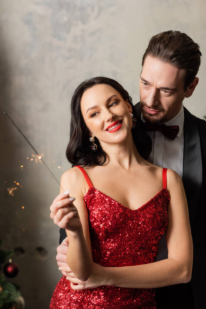 красивая богатая пара в формальной одежде улыбается и смотрит на яркие искры в канун Рождества - Фото, изображение