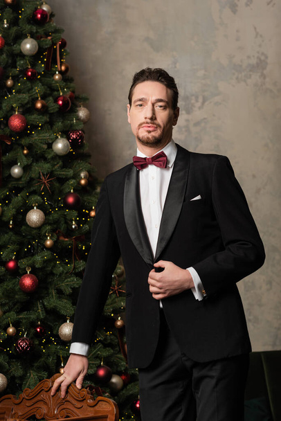 rijke heer in formele kleding met strikje kijken naar camera in de buurt van versierde kerstboom - Foto, afbeelding