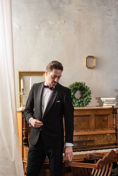 wealthy gentleman in formal attire with bow tie standing near piano and Christmas wreath on wall - Photo, Image