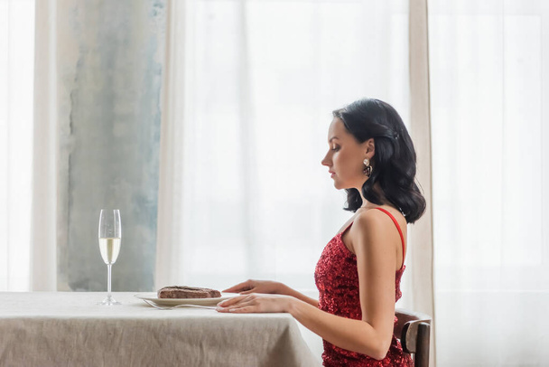 boční pohled na elegantní žena v červených šatech sedí u jídelního stolu se sklenkou šampaňského a steak - Fotografie, Obrázek