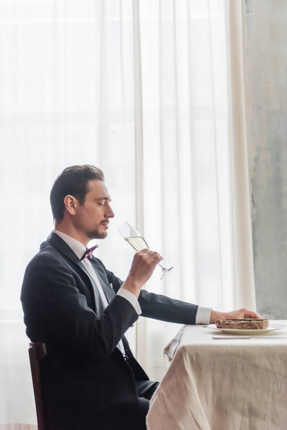 hombre guapo en esmoquin disfrutando del sabor del champán y sentado a la mesa con carne de res en el plato - Foto, imagen