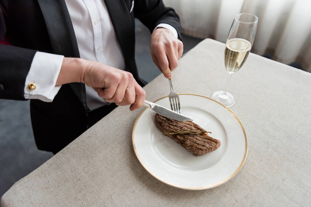 felső kilátás gazdag ember szmokingban vágás finom marha steak tányéron közelében üveg pezsgő - Fotó, kép