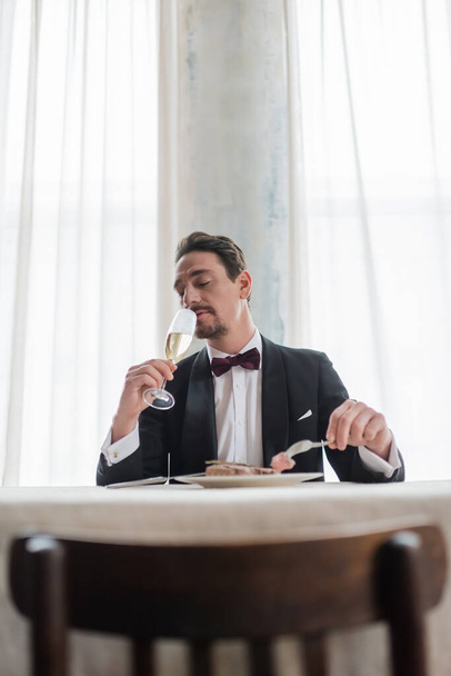 caballero bien vestido en esmoquin disfrutando del sabor del champán mientras come carne de res, la vida rica - Foto, Imagen
