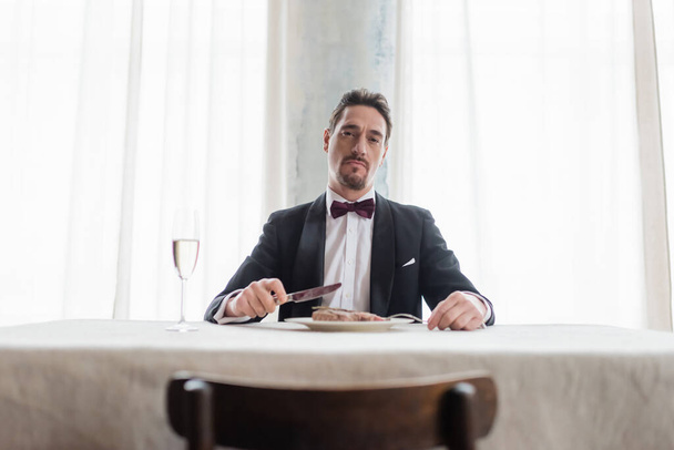 богатый джентльмен в смокинге ест вкусный бифштекс рядом с бокалом шампанского, смотрите в камеру - Фото, изображение