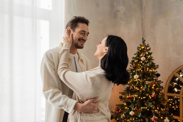 joyful couple in comfortable home wear smiling and hugging near decorated Christmas tree in bedroom - Photo, Image