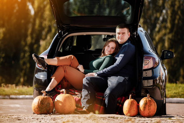 Couple in love, is sitting and hugging in car trunk at park with pumpkins decorated, on autumn warm day. Positivity and romantic man and woman celebrating thanksgiving holiday, Halloween. Fall time. - Photo, Image
