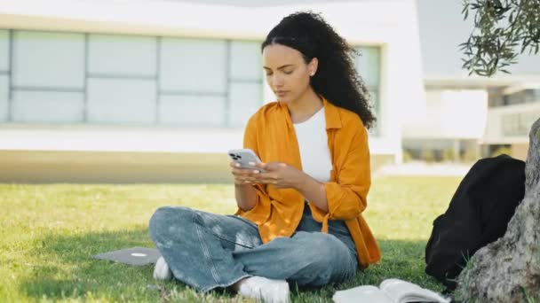 Vídeo completo de uma estudante brasileira ou hispânica frustrada, sentada na grama perto da árvore, usando telefone celular, mensagens de texto nas mídias sociais com amigos, preocupada com notícias, estressada - Filmagem, Vídeo