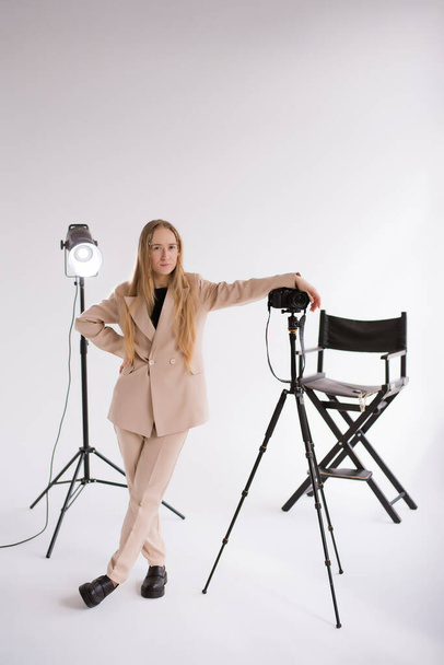 Женщина-продюсер, снимает блондинку, работающую с камерой Lumix GH5, светящуюся на штативе в студии. Ношение формального обнаженного брючного костюма на белом изолированном фоне, вертикальное  - Фото, изображение