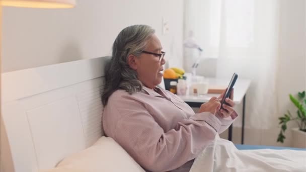 Vista lateral de mujer asiática madura charlando con sus amigos en el teléfono móvil mientras está acostada en la cama en pijama - Imágenes, Vídeo