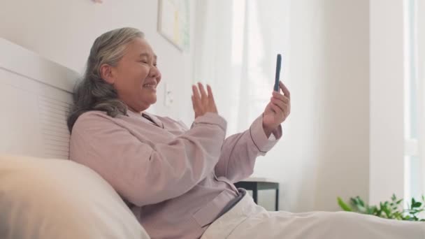 Vista lateral de una mujer asiática mayor hablando con amigos en una videollamada usando un teléfono inteligente mientras descansa en la cama - Imágenes, Vídeo