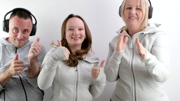 Kulaklıklı neşeli bir aile beyaz eşofmanların arka planında baş parmak göstererek dans ediyor eğlenceli aile sporları yapıyor mutlu aile anne kız baba. Neşeli müzik - Fotoğraf, Görsel