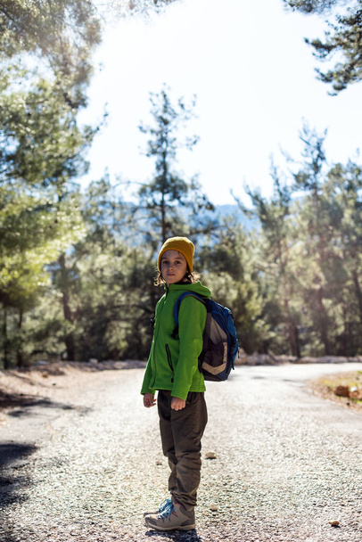 Мальчик с рюкзаком посреди лесной дороги, ребенок исследует дикую природу, ребенок стоит один среди деревьев, портрет мальчика. Путешествия и походы. - Фото, изображение