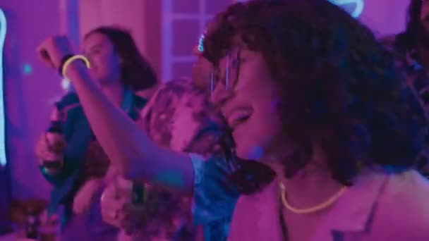 Plan rapproché moyen de jeunes filles et de jeunes hommes dansant énergiquement et s'amusant ensemble à la fête de la maison, dans la lumière bleue et rose néon, tenant des bouteilles de bière et portant des lunettes amusantes - Séquence, vidéo