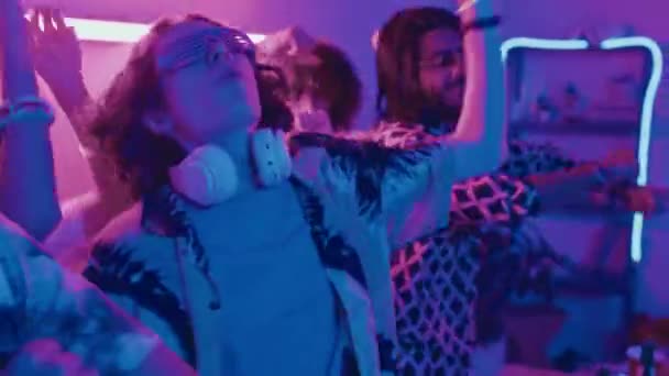 Średnie zbliżenie ujęcie szczęśliwych młodych studentów w strojach imprezowych tańczących entuzjastycznie i machających rękami, w niebieskich i różowych neonach, bawiących się w domu rave - Materiał filmowy, wideo
