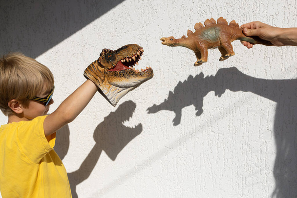 garçon et maman jouent avec le jouet dinosaure, créant des ombres contrastées intéressantes de celui-ci sur le mur. Belle journée ensoleillée. joyeuse enfance, idées de jeux, mode de vie, développement de l'imagination - Photo, image