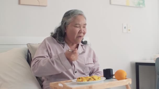 Mittlere Aufnahme einer älteren kranken Frau im Pyjama, die zu Hause auf dem Bett sitzt und das Frühstück vom Tablett aus isst - Filmmaterial, Video