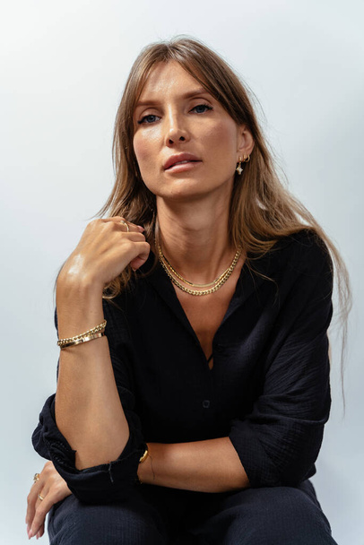 Verticaal portret van aantrekkelijke volwassen vrouw in zwart model met gouden armbanden, ringen, kettingen op witte achtergrond. - Foto, afbeelding