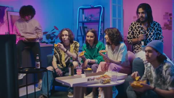 Full shot of six diverse young male and female friends in bright casual clothes hanging out together at home, watching TV, drinking beer, eating pizza and snacks, pointing at screen and talking - Footage, Video