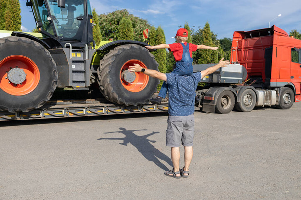muž a chlapec stojí před velkým traktorem stojícím na přívěsu, zadní pohled. Synova vášeň pro auta, táta a dítě se se zájmem dívají na auta na parkovišti. Den otců, buďte spolu - Fotografie, Obrázek