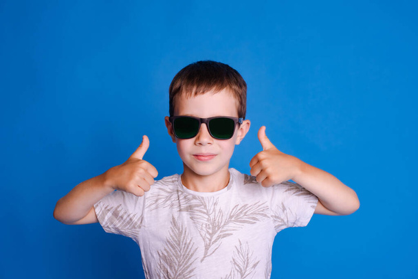 προστασία των ματιών από τον ήλιο. παιδί αγόρι σε γυαλιά ηλίου δείχνουν τον αντίχειρα πάνω από το γαλάζιο φόντο στούντιο - Φωτογραφία, εικόνα