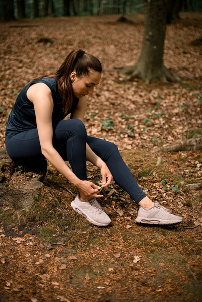 Woman runner sits on tree stump and tying her shoelaces, ready to run through forest. Active healthy lifestyle. High quality photo - Photo, Image