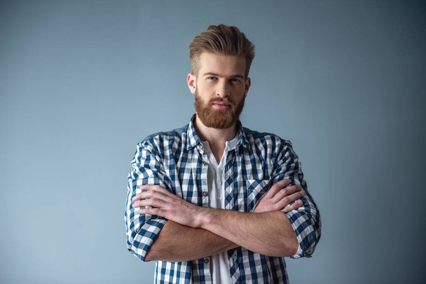 Handsome bearded man in casual clothes is looking at camera while standing with folded arms on gray background - Photo, Image