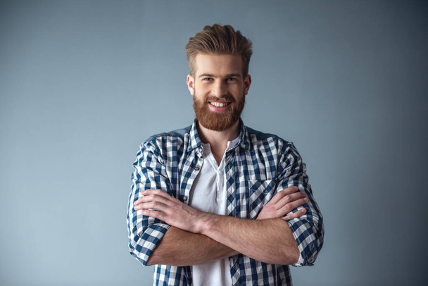 Hombre barbudo guapo en ropa casual está mirando a la cámara y sonriendo mientras está de pie con los brazos cruzados sobre fondo gris
 - Foto, Imagen