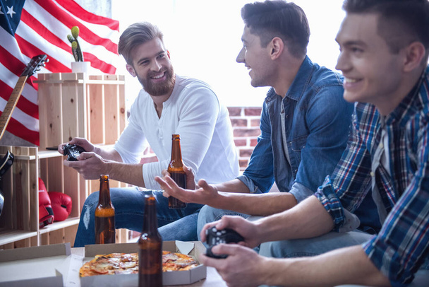 attraktive Kerle spielen Videospiele, trinken Bier, reden und lächeln, während sie zu Hause auf der Couch sitzen - Foto, Bild