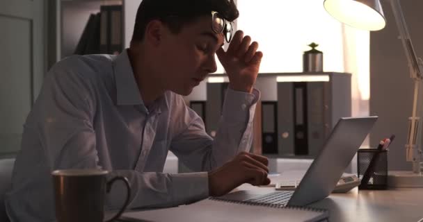 Uitgeputte kantoormedewerker zit aan het bureau, zichtbaar vermoeid van de eisen van de dag. Met een zucht van vermoeidheid wrijft de arbeider in hun ogen, op zoek naar verlichting van de druk veroorzaakt door lange werkuren.. - Video