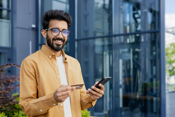 Portrait of young man outside office building, hispanic smiling and looking at camera, holding phone and bank credit card, online shopping and booking services remotely. - Fotografie, Obrázek