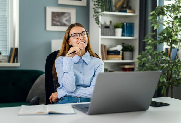 Young thoughtful woman wearing eyeglasses dreaming at desk with laptop, motivated idea concept, smiling female working in cozy light room - Foto, Imagem