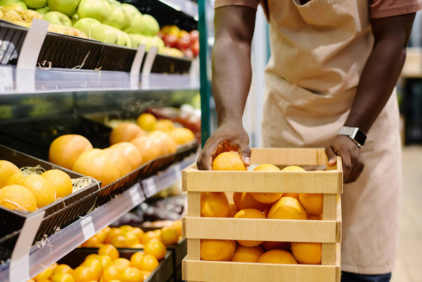 Verkäuferin in Uniform legt Obst aus Schachtel in Regalen aus - Foto, Bild