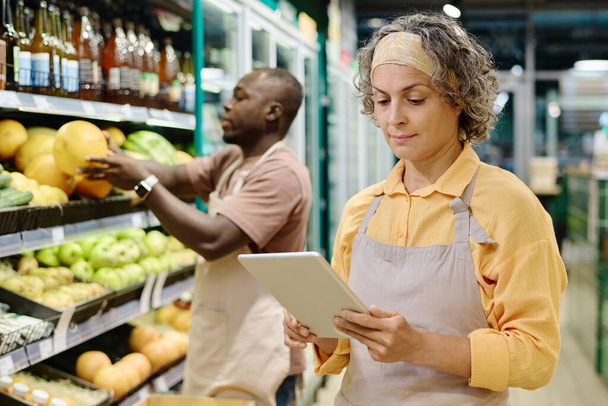 Verkäuferin in Uniform steuert Waren online auf Tablet-PC während ihrer Arbeit im Supermarkt - Foto, Bild