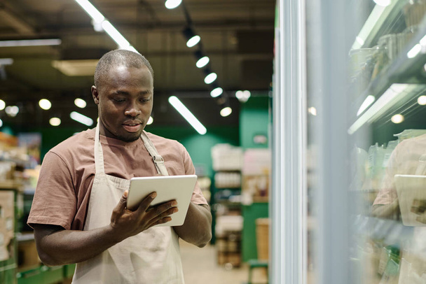 Afrikai-amerikai szupermarket munkavállaló egyenruhában használja tabletta pc a munkáját, hogy ellenőrizzék eltarthatóságát áruk online - Fotó, kép