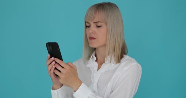Woman with a bright smile while texting on her phone against a serene blue background. Her cheerful expression and engagement with the device suggest a positive and enjoyable conversation. - Footage, Video