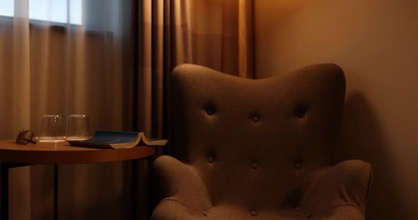 Bruine bank en houten salontafel met boek en leesbril in semi-donkere hotelkamer. Plaats voor ontspanning en leesboek sessie thuis - Video