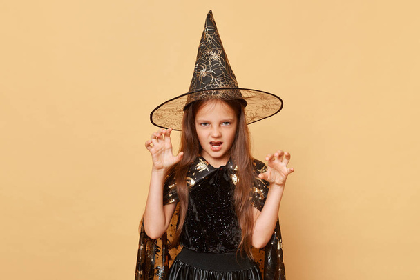 Spooky celebration. Little witch. Childhood sorcery. Carnival fun. Scary female child in Halloween costume and witch hat isolated over beige background raised arms frighting guests and halloween party - Φωτογραφία, εικόνα