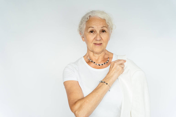 Ευτυχισμένο πορτρέτο ηλικιωμένη γκρίζα μαλλιά γυναίκα φορώντας λευκά ρούχα δείχνει κοσμήματα bijouterie σε λευκό φόντο. - Φωτογραφία, εικόνα