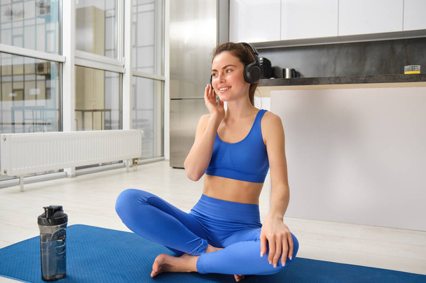 Young sporty athletic fitness trainer instructor woman wear blue tracksuit spend time in home gym use mobile phone headphones listen music. Workout sport concept. - Photo, Image