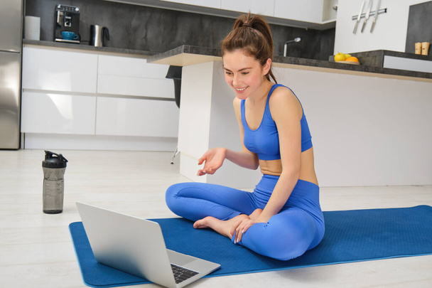 Image of young fitness instructor using laptop for online workout training, showing fitness exercises, talking to client, sitting on yoga rubber mat in sportswear. - Photo, Image