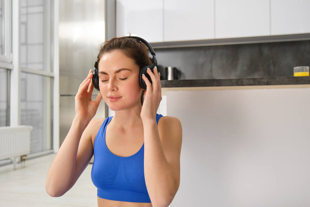 Mujer joven se pone auriculares inalámbricos para escuchar música durante el entrenamiento en casa, lleva sportsbra, sonríe con la cara complacida. - Foto, imagen