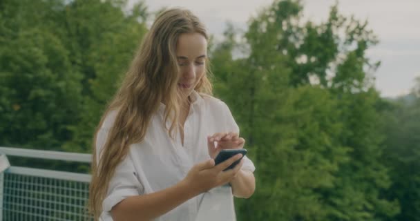 Αργή κίνηση πλάνο χαμογελαστή ξανθιά γυναίκα κρατώντας έξυπνο τηλέφωνο, ενώ στέκεται κοντά κιγκλίδωμα στα δέντρα - Πλάνα, βίντεο