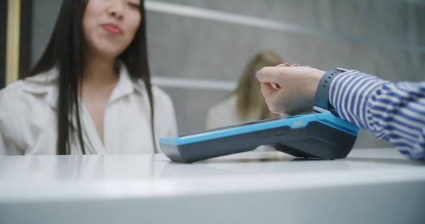 Asian female administrator stands at reception desk in clinic, gives terminal. Woman pays with contactless payment for appointment with doctor using watch. Medical staff work in modern medical center. - Photo, Image
