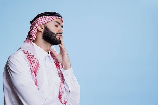 Bedachtzame moslim man met traditionele kleren die de kin aanraken terwijl hij poseert in de studio met een penseelachtige uitdrukking. Verbijsterd arabisch persoon in thobe en hoofdtooi denken over oplossing - Foto, afbeelding