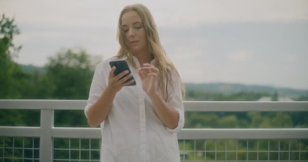 Αργή κίνηση shot της νεαρής ελκυστική γυναίκα χρησιμοποιώντας έξυπνο τηλέφωνο, ενώ ακουμπά στο κιγκλίδωμα στον ουρανό - Πλάνα, βίντεο