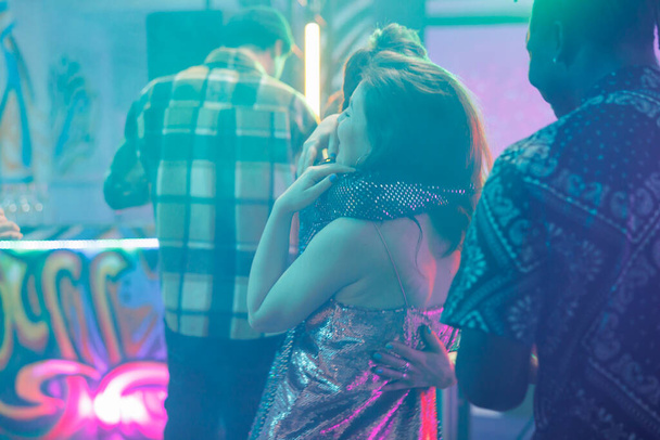 Women hugging in nightclub while attending discotheque party. Girlfriends embracing on dancefloor with spotlights while dancing and enjoying nightlife entertainment in club at night - Photo, Image